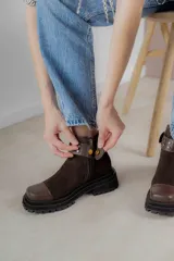 Bota corta de cuero gamuzado color marrón chocolate, con punta cuadrada y suela track. Presenta una correa con hebilla desmontable y cierre lateral con cremallera.