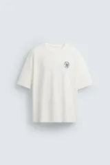 Camiseta blanca de corte holgado, cuello redondo y mangas cortas. Presenta un bordado y estampado combinado en el frente.