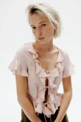 Blusa color rosa pálido de lino y viscosa, con escote en V pronunciado, mangas cortas y volados en el escote, mangas y ruedo. Se ata en el centro con dos cordones finos.