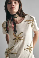 Remera de mangas cortas color crudo, de calce relajado y escote redondo. El frente se encuentra bordado con un diseño de palmeras en lentejuelas y canutillos.