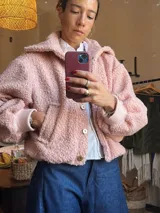 Chaqueta corta color rosa con textura de lana de oveja, cuello camisero, cierre frontal con botones dorados, bolsillos laterales y puños tejidos.