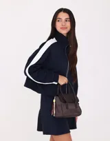 Cartera tipo city de tela deportiva color marrón con doble asa corta de mano con velcro, correa regulable desmontable, solapa con broche con botón para cerrado y charm colgante.