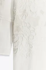 Vestido corto de algodón y lino color blanco, con cuello babero cuadrado, manga larga y bordados florales. Cierre en la espalda con abertura y botón.