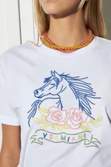 Remera blanca de algodón con cuello redondo y mangas cortas. Estampado frontal con un caballo azul, rosas rosadas y texto bordado en hilo amarillo.