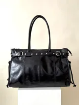 Bolso grande de cuero negro con estructura rígida y asas de hombro. Presenta detalles de tiras con tachas y hebillas en los extremos superiores, y lazos ajustables en los laterales. El interior no tiene forro y cuenta con un bolsillo tipo sobre con cierre.