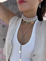 Gargantilla choker con hilo trenzado color blanco, adornada con conchas marinas tipo buzios y medallitas bañadas en oro y acero quirúrgico. Se cierra con cadena de acero quirúrgico y ganchito para ajustar el largo.