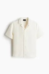 Camisa de manga corta color blanco hueso con textura. Tiene cuello abierto, tapeta lisa y bajo recto.