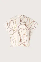 Camisa blanca con estampado de cadenas y cuerdas en tonos dorados y marrones. Tiene cuello mao, mangas cortas y detalles de encaje en el frente y la espalda.