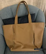 Bolso tipo tote de cuero color beige con textura granulada y tachas plateadas en los laterales.