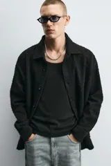 Camisa negra de denim de algodón con textura irregular, corte relaxed fit, cuello solapa, manga larga con puños abotonados y cierre frontal con botones.
