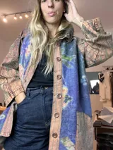 Campera larga con cuello, confeccionada con retazos de tela. El cuerpo principal es de tejido afelpado azul con estampado floral en tonos amarillos y verdes. Los bordes, solapas y mangas son de tela con estampado a rayas en tonos terracota y marrón. Cierre frontal con botones marrones.