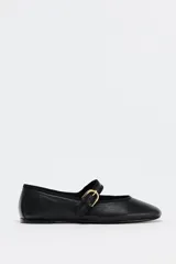 Zapato plano tipo bailarina color negro, confeccionado en piel, con tira delantera con hebilla metálica ajustable y acabado en punta redonda.