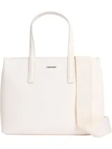 Cartera tipo tote pequeña de color blanco, con asas de mano fijas y correa ajustable opcional. Presenta un logo metálico de Calvin Klein en la parte delantera y un logo bordado en la correa de tela.