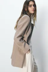 Abrigo color beige confeccionado en lana. Tiene cuello subido, mangas largas con hombreras, forro interior combinado y cierre frontal cruzado con botones metálicos.