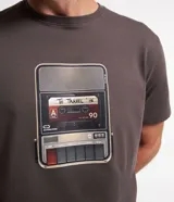 Remera gris de algodón con cuello redondo y mangas cortas. Presenta un estampado frontal de un reproductor de casete con la leyenda "To Travel".