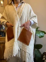 Ruana color crema tejida en lana merino, con flecos y bolsillos de cuero marrón.