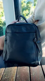 Mochila negra de símil cuero con costuras diagonales en el frente. Tiene un bolsillo frontal con broche magnético, asa de mano y correas regulables.