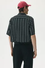 Camisa de manga corta con rayas verticales blancas sobre fondo negro, cuello inglés, tapeta con botones y bolsillo de parche en el pecho. Corte recto y holgado.