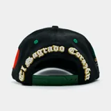 Gorra de béisbol negra con visera estampada con diseño paisley verde y blanco. Presenta un bordado de la Virgen de Guadalupe en el panel lateral y una letra 'M' estilizada en el frente con franjas blancas, verdes y doradas.