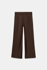 Pantalón marrón de tiro alto con bolsillos laterales ocultos en costuras, pliegues en delantero y cinturón doble en mismo tejido. Cierre frontal con cremallera y gancho metálico.