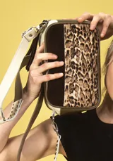 Cartera bandolera de cuero color arena con frente de pelo con estampado de leopardo, laterales de gamuza color chocolate y bordes de cuero verde oliva. Tiene correa ancha regulable con tachas metálicas y doble cierre superior independiente.