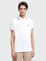 Polo de piqué de algodón blanco de manga corta, con cuello y puños con ribetes tricolor (rojo, blanco y azul marino). Presenta un pequeño logo bordado en el pecho.