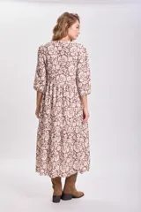 Vestido largo con estampado floral en tonos mostaza, marrón y blanco, con mangas largas abullonadas y falda con paneles. Cinturón marrón con tachas.