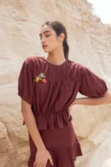 Blusa color chocolate con cuello redondo, mangas cortas abullonadas, detalle de flores bordadas en el frente y corte con volantes en la cintura.