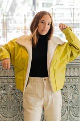 Chaqueta bomber color amarillo de cuero sintético con interior de piel sintética color crema, cierre frontal con cremallera, corte a la cintura, hombros caídos y mangas amplias.