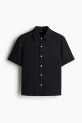 Camisa negra de punto con textura, de corte recto, manga corta, cuello inglés, tapeta lisa, canesú en la espalda y bajo recto con aberturas laterales.