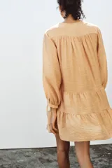 Vestido corto color beige con cuello redondo, cordón con borlas y manga larga. Presenta bordados combinados a contraste en la pechera, cierre frontal con botones ocultos, forro interior y bajo con volantes.