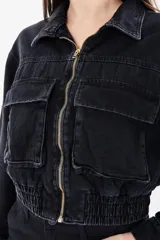 Chaqueta corta de denim negro con cuello clásico, cierre frontal con cremallera metálica, dos bolsillos de parche con solapa en el pecho y puños con botón.