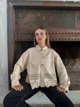Camisa color beige con cuello clásico, cierre frontal con botones dorados, bolsillos de parche en el pecho y detalle de calado con terminación festoneada en el ruedo.