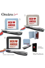 Billetera tipo monedero de color celeste claro, con cierre de cremallera y detalle frontal con texto 'MON AMOUR' en un parche blanco. Incluye un llavero desmontable con forma de labios rojos y una correa de mano negra con franjas blancas.