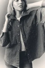 Camisa de denim azul con apliques metálicos plateados, de calce amplio, cuello camisero, cierre frontal con botones a presión, mangas largas con puños abotonados y dos bolsillos con solapa abotonada en el pecho.