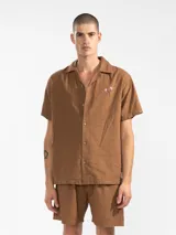 Camisa marrón de lino y algodón, de corte holgado, con cuello tipo camp y cierre frontal con botones de madera. Presenta un bordado de palmeras en el pecho izquierdo.