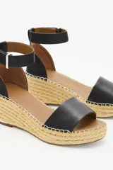 Sandalia de cuero vacuno color negro con taco tipo cuña revestido en yute. Presenta una tira ancha sobre el empeine y una pulsera ajustable al tobillo con cierre de hebilla metálica.