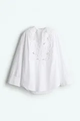Blusa blanca de algodón con escote en V y pechera con broderie tono sobre tono embellecido con minicuentas y ribetes con pompones. Modelo de corte holgado con hombros caídos y mangas largas con tapeta de manga y puños abotonados. Bajo redondeado.