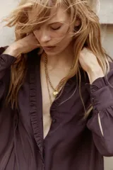 Vestido corto color marrón oscuro, estilo camisero, con cuello mao con ribete de volado y tapeta de botones con el mismo detalle. Posee mangas largas con puños sencillos de volado y un cinturón fino extraíble para ajustar la cintura. La falda tiene un fruncido escalonado que le da vuelo.