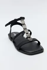 Sandalias planas color beige con tiras finas y aplique metálico central. Cierre con hebilla lateral y base marrón.