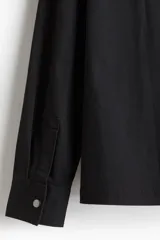 Sobrecamisa negra de algodón con cuello inglés, cierre bidireccional, bolsillos superiores con solapa y botones a presión, canesú en la espalda, bajo recto con aberturas laterales y mangas largas con tapeta de manga y botón a presión en los puños.