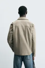 Sobrecamisa color arena de corte recto, con cuello solapa, cierre frontal con cremallera, manga larga con puños abotonados, dos bolsillos de plastrón con solapa en el pecho y dos bolsillos laterales en la costura de la cadera.