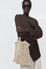 Bolso tipo shopper de tamaño pequeño, tejido en crochet color beige claro con patrón calado. Incluye asas de hombro de cuero sintético marrón oscuro y una bolsa interior con cierre de cremallera.