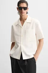 Camisa color blanco con textura de rayas verticales, corte relaxed fit, cuello bowling, manga corta, bolsillo de parche en el pecho y cierre frontal con botones.
