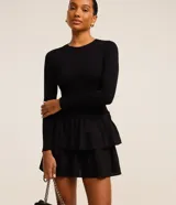 Vestido corto negro de viscosa con mangas largas, cuello redondo y falda godê con superposición de volantes. La parte superior es acanalada y la inferior tiene capas fruncidas.