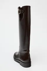 Bota alta de cuero color marrón oscuro, con caña alta, punta redonda y hebilla decorativa en la parte superior.