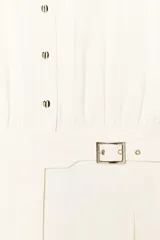 Mono largo color crema, de cuello solapa y manga corta con dobladillo. Tiene bolsillos delanteros y falsos bolsillos traseros. El pantalón es de pierna ancha con pinzas delanteras y dobladillo. Se cierra con cremallera, hebilla y botones.