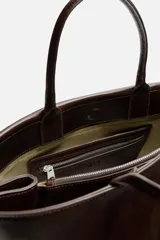 Bolso de mano extragrande color marrón oscuro, fabricado en piel de alta calidad. Tiene un compartimento principal con cierre magnético, un bolsillo interior con cremallera y un bolsillo secundario con solapa y cinta. Las asas son de piel.
