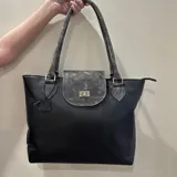 Cartera tipo tote de cuero negro con detalles en gris. Tiene cierre con tapa y broche metálico, doble asa de hombro y un dije colgante con forma de oso.