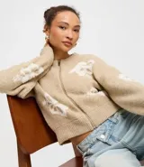 Cardigan cropped color beige con estampado de flores blancas, cuello redondo, cierre frontal con cremallera y mangas largas.